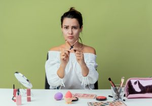 annoyed beautiful girl sits table with makeup tools crosses makeup brushes gesturing no isolated green wall