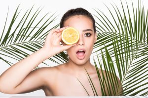 young woman with perfect skin holding citrus fruit hand surrounded by palms leaves isolated white wall