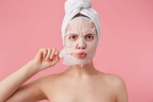 close up frowning young woman with towel her head after shower trying remove fabric mask from face stands